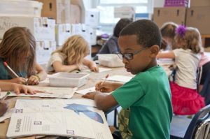 A kid is at the forefront looking at the book in front of him while using modern teaching aids to learn. Other kids are in the background.